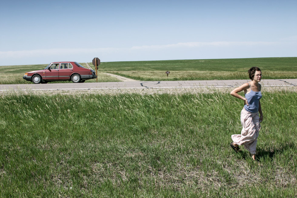 Photograph of a woman walking on a green prairie with a red car and road in the distance.